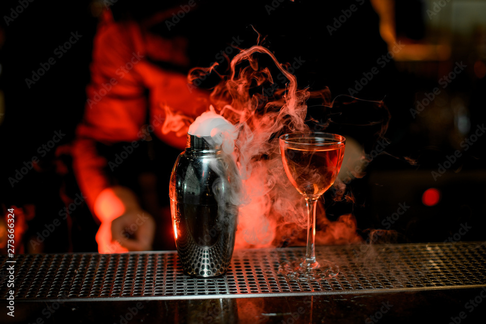 Bartender standing in the foreground of cocktail in the glass and smoky steel shaker