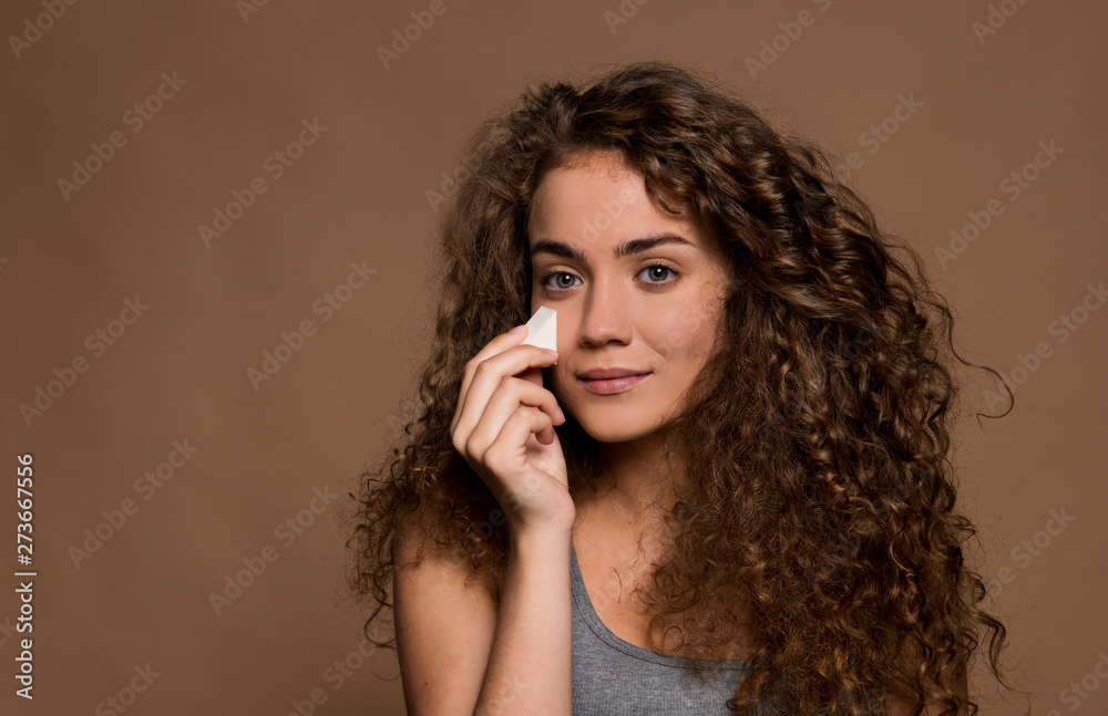 Fototapeta premium Portrait of a young woman applying make-up in a studio against brown background.