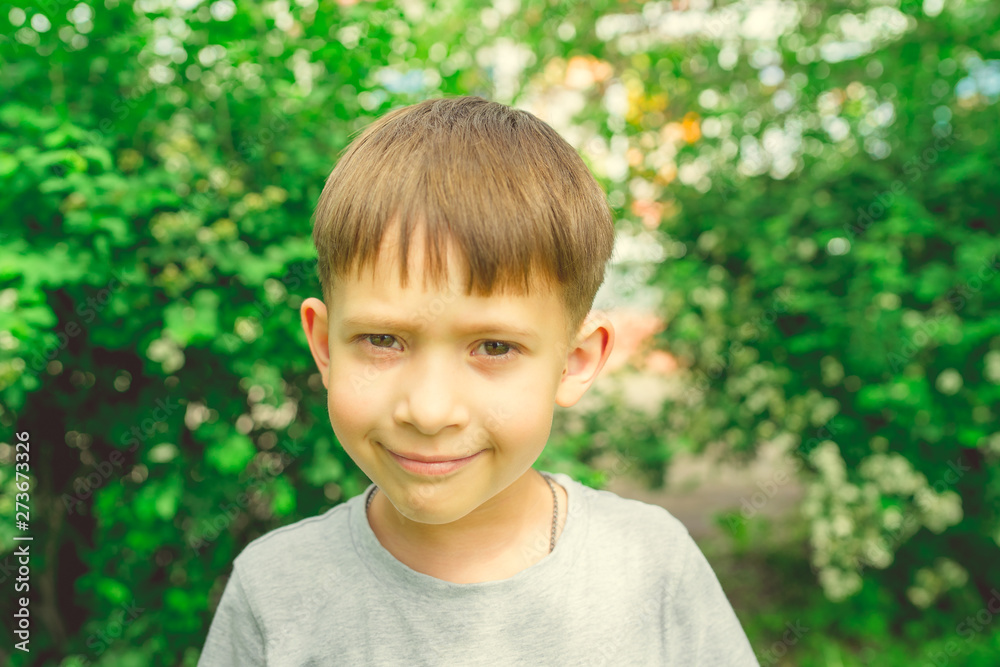 Serious boy in bright green garden. Adorable cute kid with stylish hair dress standing in bright greenery in street Serious boy in bright green garden. Adorable cute kid with stylish hair dress standing in bright greenery in street