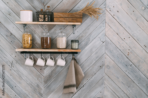Cereals in glass jars on shelf, white cups, towel hang on hooks on kitchen wall