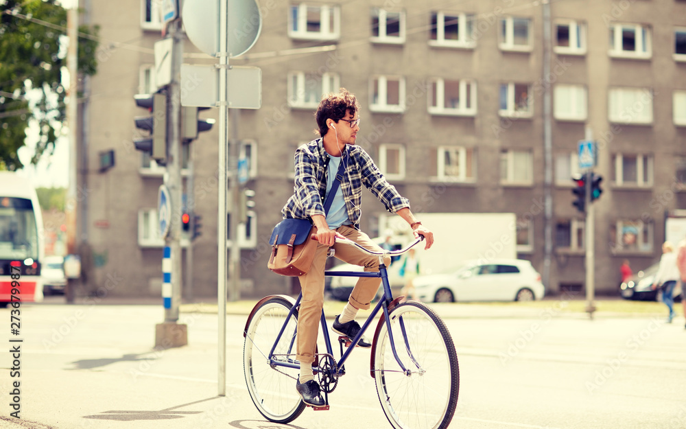 Obraz premium people, style, leisure and lifestyle - young hipster man with shoulder bag and earphones riding fixed gear bike on city street
