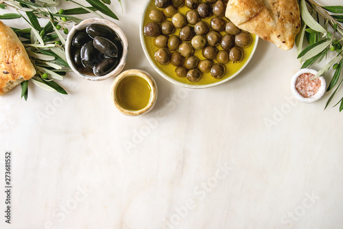 Variety of green and black whole olives in olive oil served in ceramic bowls with fresh baked ciabatta bread, pink salt and young olive wood branches over white marble background. Flat lay, copy space photo