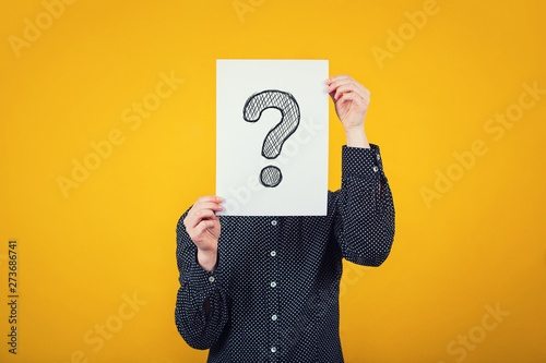 Businesswoman covering face using a white paper sheet with drawn question mark, like a mask, for hiding her identity. Isolated on yellow wall background photo