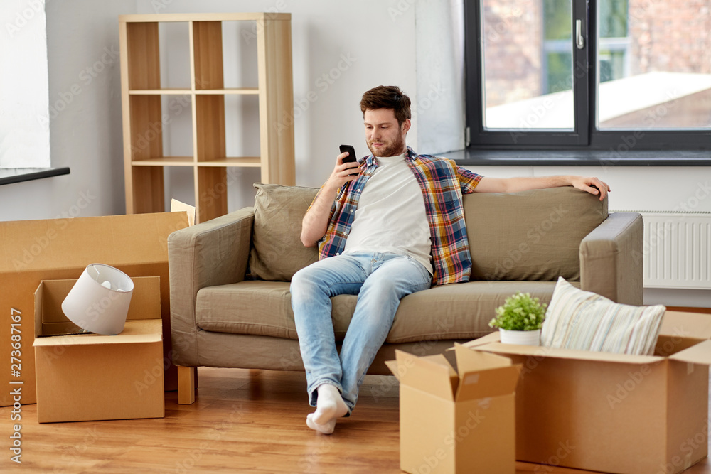 moving, people and real estate concept - happy man with smartphone and boxes sitting on sofa at new home moving, people and real estate concept - happy man with smartphone and boxes sitting on sofa at new home