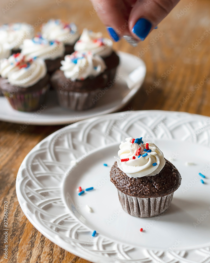 Chocolate mini cupcake on white plate being decorated with red, white, and blue sprinkles. Fingers with motion blurr in photo. Additional decorated cupcakes blurred in background. Chocolate mini cupcake on white plate being decorated with red, white, and blue sprinkles. Fingers with motion blurr in photo. Additional decorated cupcakes blurred in background.