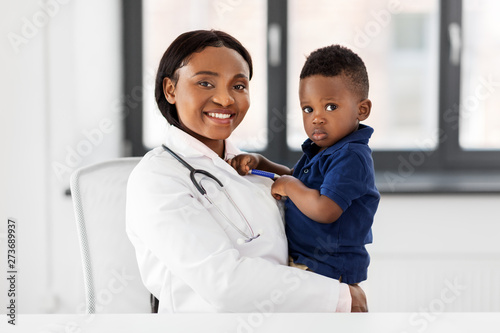 medicine, healtcare, pediatry and people concept - happy african american female doctor or pediatrician holding baby patient on medical exam at clinic