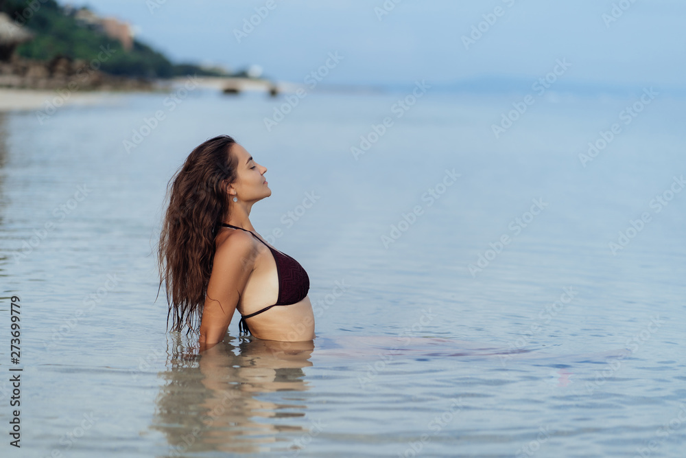 Naklejka premium Young tanned girl in swimsuit swims in ocean and poses on beach at vacation