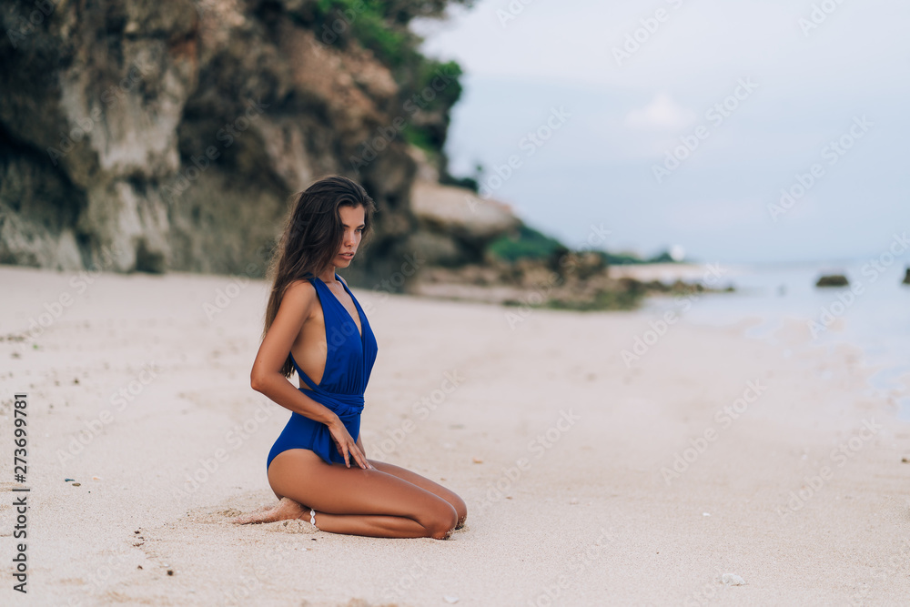 Beautiful sexy tanned girl in blue swimsuit sits on white sand beach  