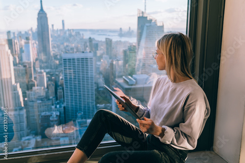 Stylish blonde female in casual wear observing scenic views of Empire State building landmark and cityscape downtown while sitting in window sill. Popular attractions of New York City. Digital nomad photo