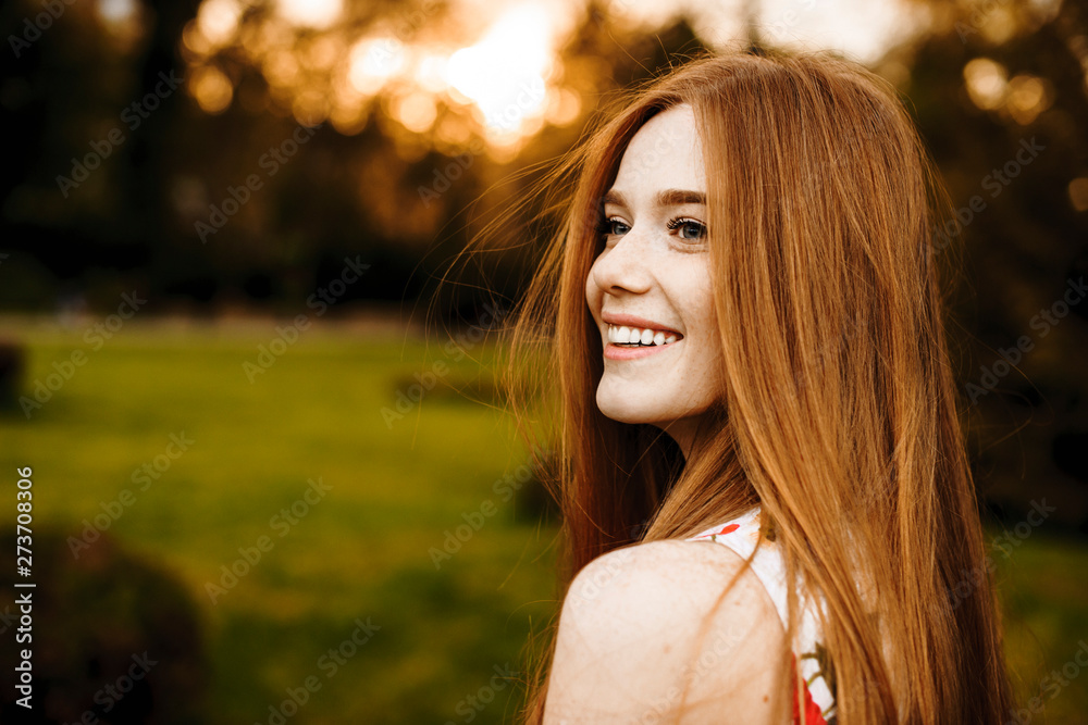 Portrait of a lovely female with long red hair and freckles looking away laughing against sunset outside. - obrazy, fototapety, plakaty 