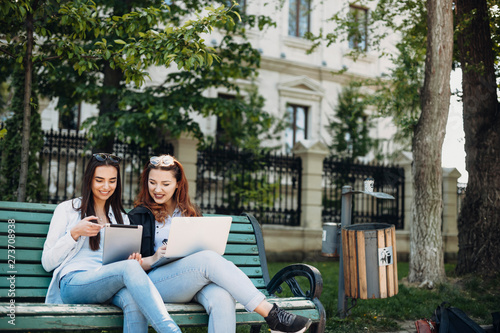 Two handsome young caucasian woman sitting on a bench smiling while looking to a tablet screen outside against a building.