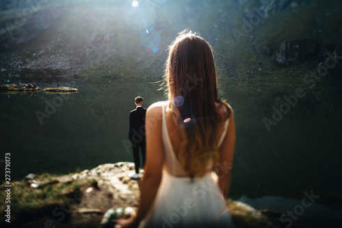 Back view of a unrecognizable groom sitting at the edge of a lake in the mountains while his bride is looking at him.
