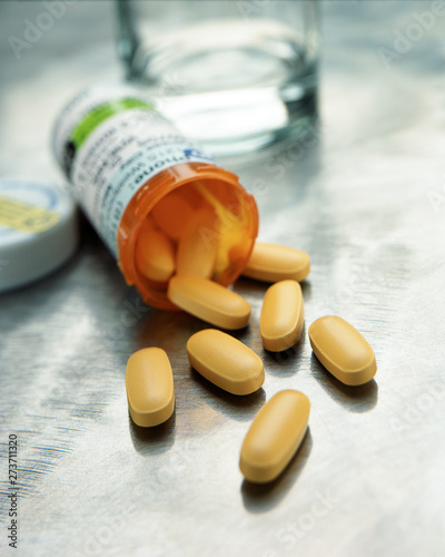 Selective focus image of yellow capsules spilling from prescription medication bottle with glass of water on steel counter photo