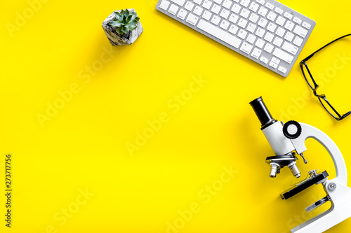 Do medical research on laboratory desk with microscope, keyboard, glasses on yellow background top view mockup