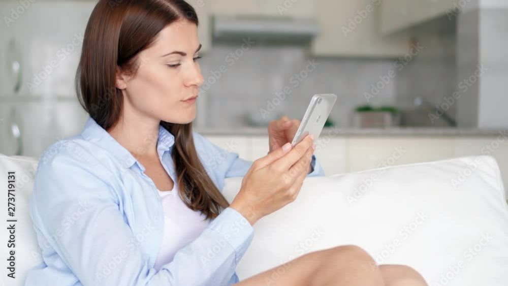 Young brunette woman with brown eyes sitting on sofa in living room using smartphone. Female housewife browsing on mobile phone chatting online with friends. Kitchen on background Young brunette woman with brown eyes sitting on sofa in living room using smartphone. Female housewife browsing on mobile phone chatting online with friends. Kitchen on background