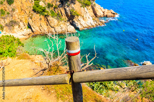 Trail post with white and red sign marking path in Tossa de Mar town with sea in background, Costa Brava, Spain