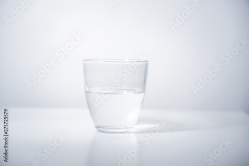 Round glass filled with cold mineral water on bright, blank surface. Reflection of the glass visible on the surface. High exposure photo.