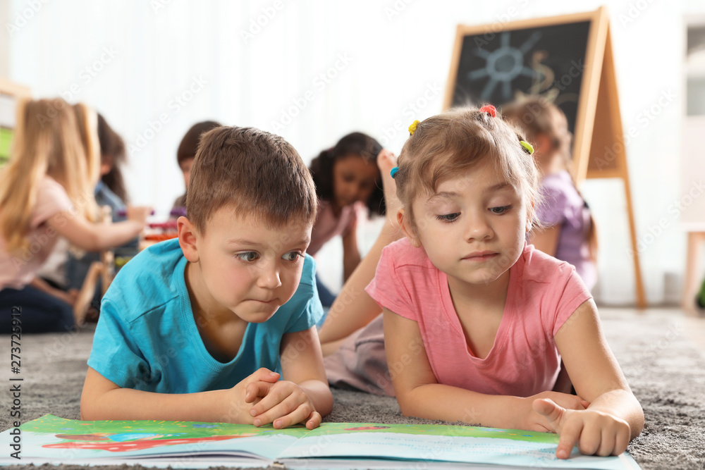 Cute kids reading book on floor while other children playing together in kindergarten Cute kids reading book on floor while other children playing together in kindergarten