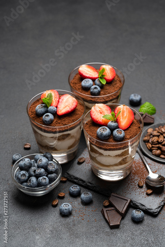 Classic tiramisu dessert with blueberries and strawberries in a glass on stone serving board on dark concrete background