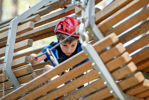 Caucasian boy enjoying activity in climbing adventure park at sunny summer day. Kid climbing in rope playground structure. Child in forest adventure park, extreme sport. © Artem