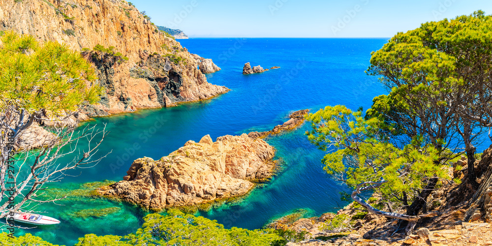 Panoramic view of white motorboat in beautiful sea cove of Cala Marquesa with green pine trees on high rock cliffs, Costa Brava, Spain