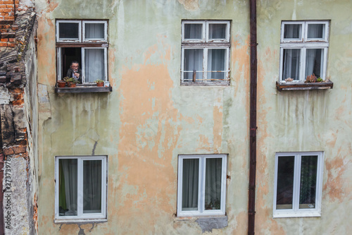 A senior man smokes in a wooden window of an old high-rise building with peeling walls. concept of past years. aging house, aging man with gray hair