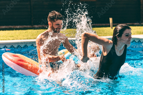 Young woman splashing on man sitting on airbed in the pool