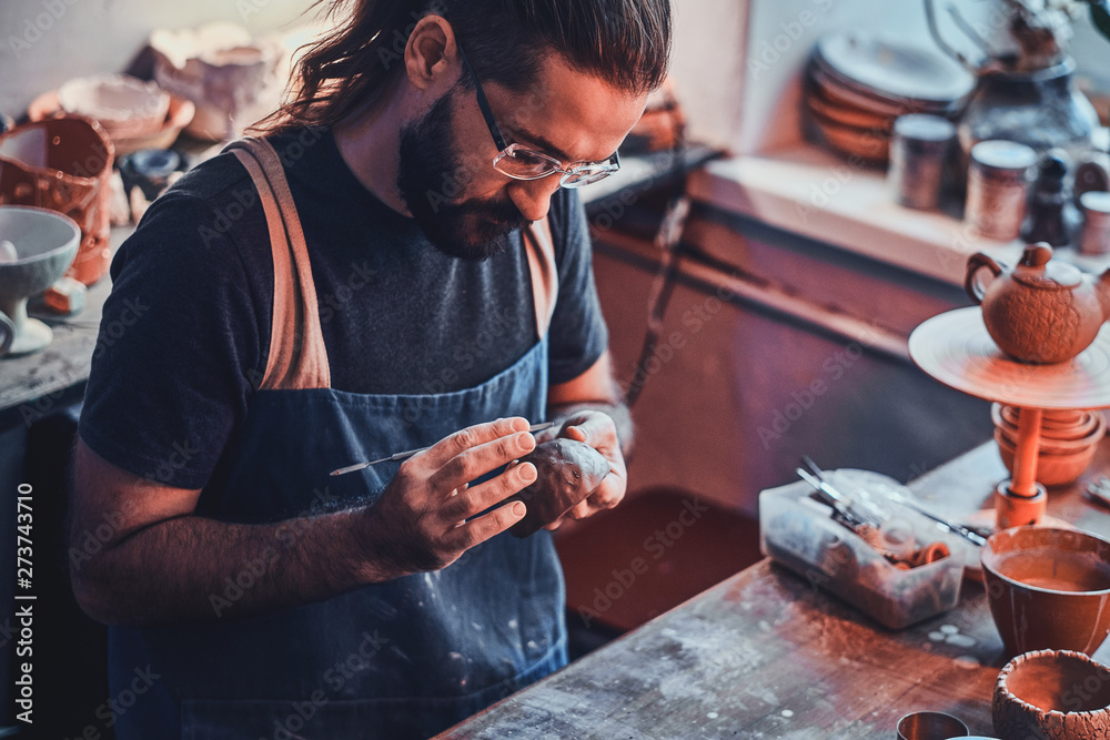 Diligent pensive man in glasses at his pottery workshop is workig for new project. - obrazy, fototapety, plakaty 