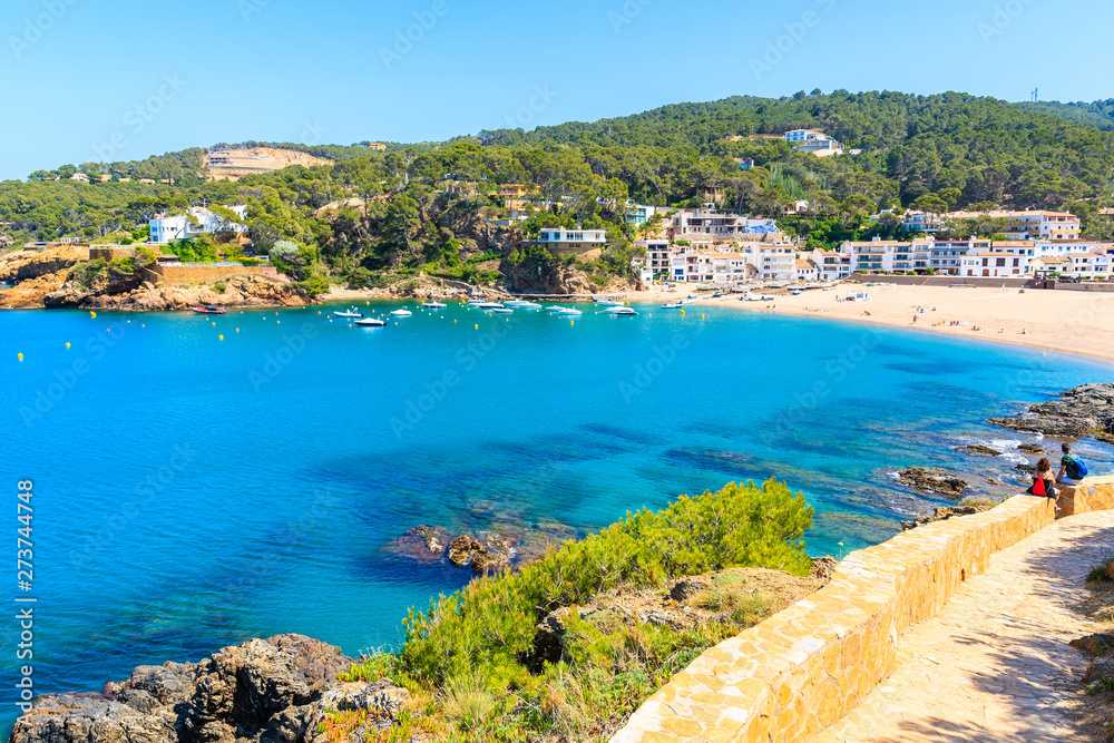 Couple of tourists sitting on coastal path and looking at beautiful bay and beach in Sa Riera village, Costa Brava, Spain