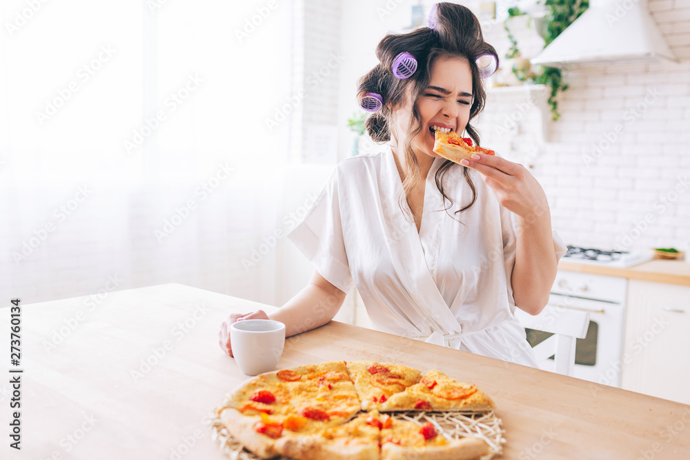 Hungry young housewife sit in kitchen and eat pizza. Bite slice of food. Alone in room. Housekeeper enjoy life without work. Hold cup in hands.