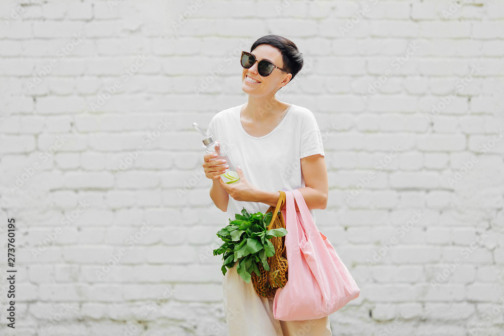 Stockfoto Young woman in light summer clothes with a eco bag of vegetables,  greens and reusable water bottle. Sustainable lifestyle. Eco friendly  concept. | Adobe Stock