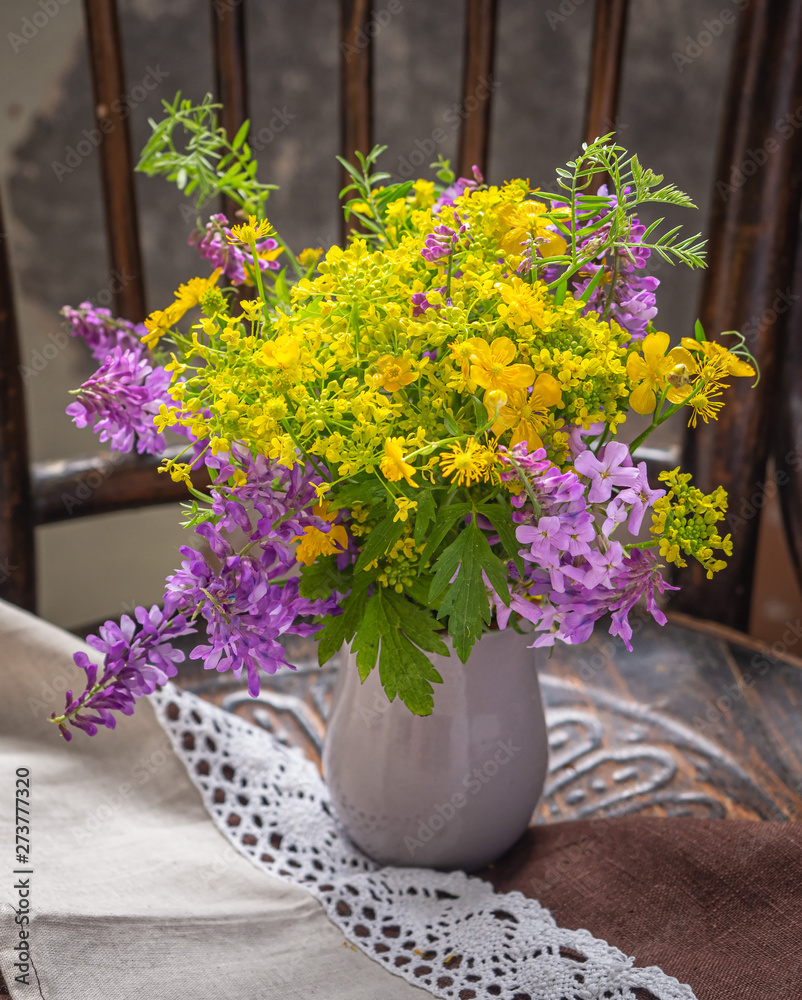 Summer bouquet of yellow and purple wild flowers in an earthenware gray vase on an old chair in a rustic interior