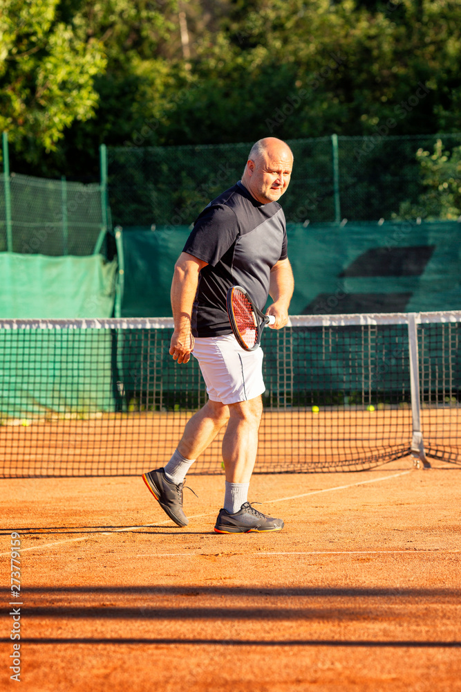 A middle-aged bald man emotionally plays tennis on the court. Loses the opponent. Outdoor. Vertical.