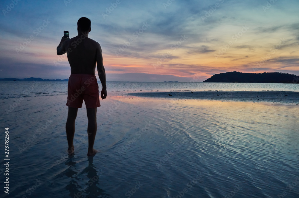 Silhouette of an athletic man standing in the sea with smart phone in hand at sunset                                  