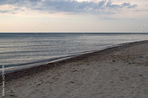 Sandy coastline covered with wavy tracks goes into perspective diagonally, azure sea with smooth surface reflecting clouds in calm weather connects with overcast sky on background of sea horizon