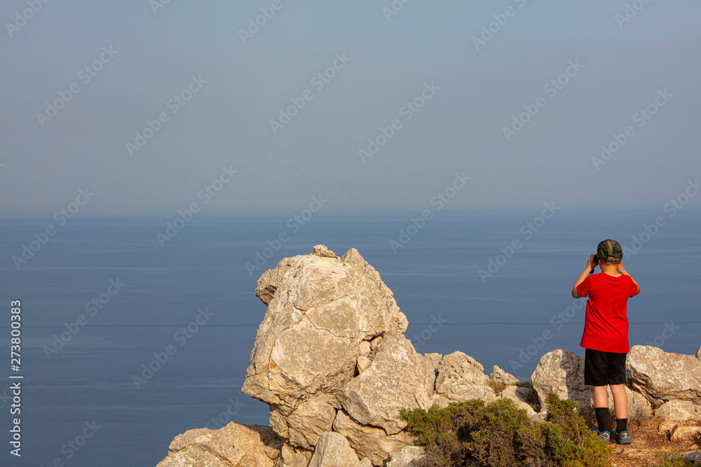 Little boy looking at binoculars on blue sea in Rhodes Greece. The child is on a high mountain.