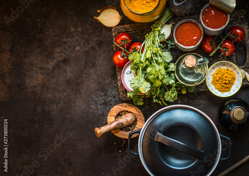Healthy ingredients for tasty tomato soup: tomatoes, fresh herbs, spices, tinned tomatoes on dark rustic kitchen table with cast iron cooking pot, top view. Copy space. Food background for your design
