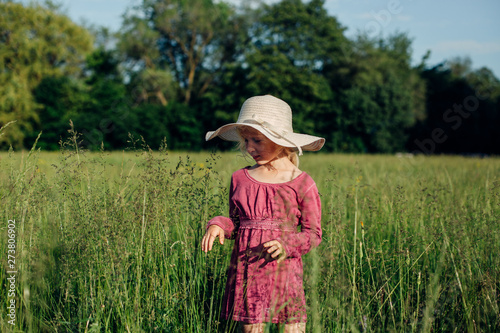 Cute preschool adorable Caucasian girl in straw hat and red pink rustic dress walking in tall high grass in meadow outdoor. Happy child kid enjoying summer. Lifestyle authentic childhood. © anoushkatoronto
