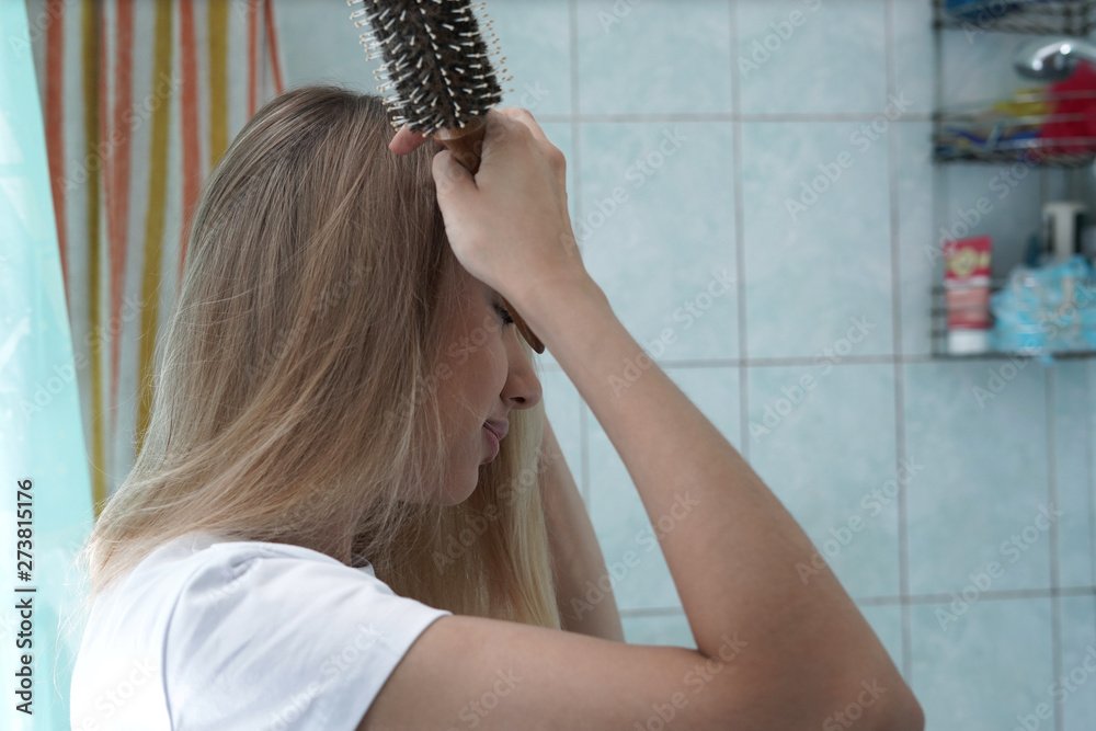 Naklejka premium Young beautiful blonde is brushing her hair in the bathroom