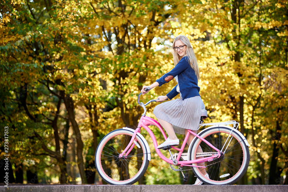 Side view of active blond long-haired attractive woman in glasses, skirt and blouse riding modern pink lady bicycle on lit by autumn sun park stairs on bright colorful golden bokeh trees background.