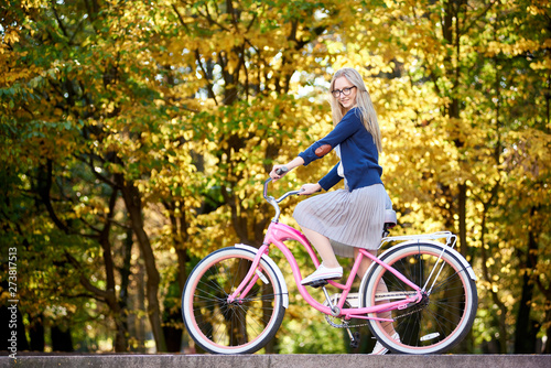 Side view of active blond long-haired attractive woman in glasses, skirt and blouse riding modern pink lady bicycle on lit by autumn sun park stairs on bright colorful golden bokeh trees background.