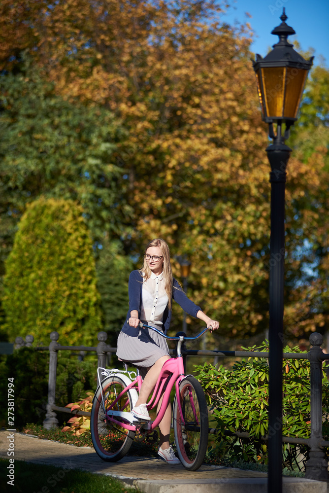Active blond long-haired attractive traveler girl in glasses and casual clothing, cycling modern pink lady bike along narrow wooden park bridge on bokeh background of beautiful different green trees. Active blond long-haired attractive traveler girl in glasses and casual clothing, cycling modern pink lady bike along narrow wooden park bridge on bokeh background of beautiful different green trees.