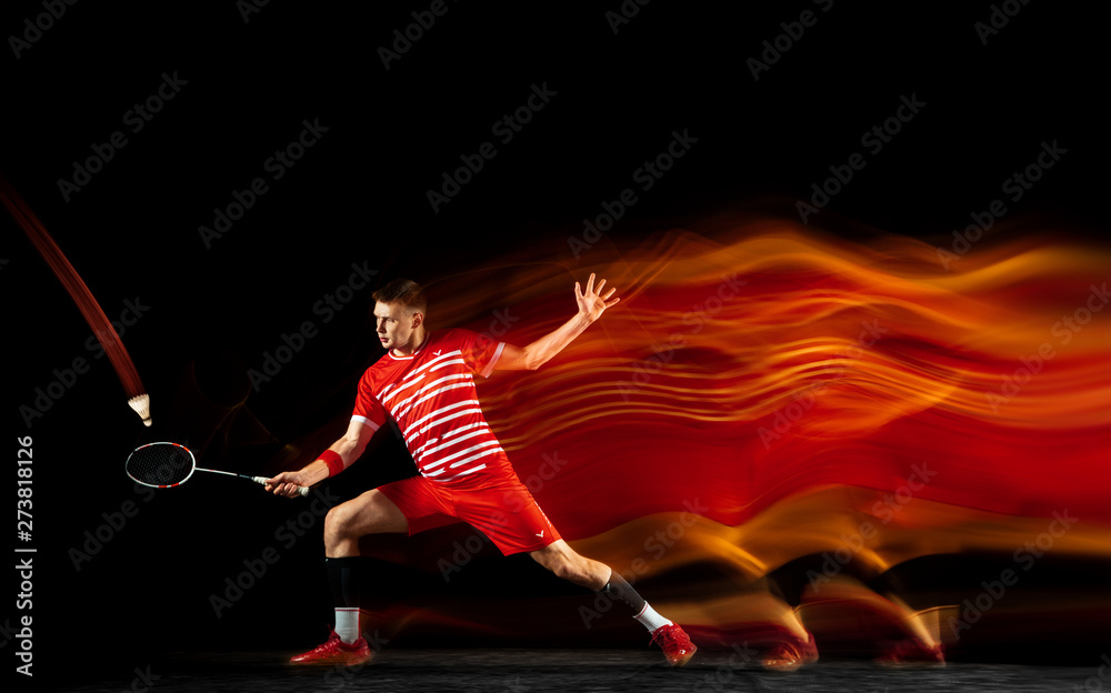 Young man playing badminton isolated on black background in mixed light.  Male model with the racket in action, motion in game with the fire shadows.  Concept of sport, movement, healthy lifestyle. Stock