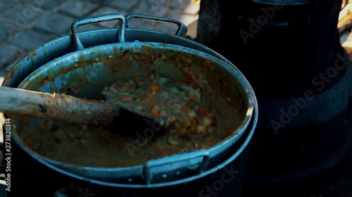 a female sewer pours soup into a clay mold bowl with a wooden spartle from a large saucepan outdoor. Medieval cusine, refugee camp, outdoor feeding concept, traditional cooking, Pirate food pottage
