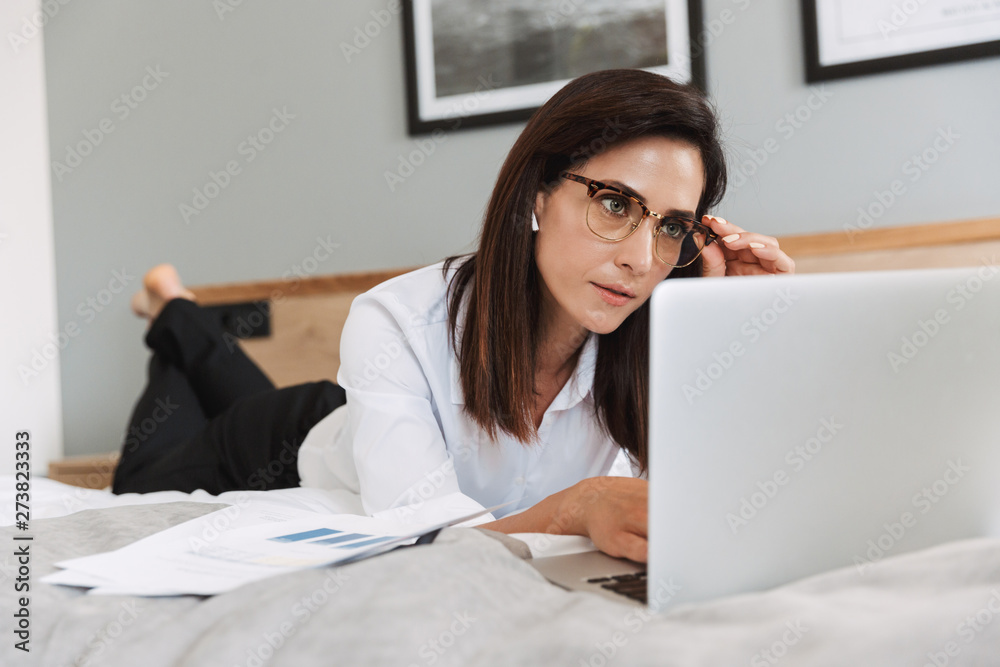 Portrait of smart adult businesswoman working with laptop and paper documents while lying on bed in apartment Portrait of smart adult businesswoman working with laptop and paper documents while lying on bed in apartment