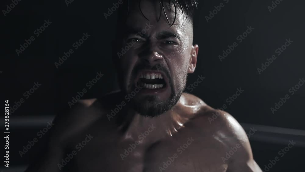 Angry man with boxing gloves screams emotionally looking ar the camera standing on the boxing ring. Slow motion.