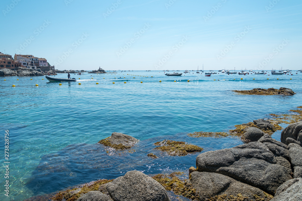 A clear and blue morning over the seashore of Calella. A boat arrives to the village among boats and the rocks at foreground.