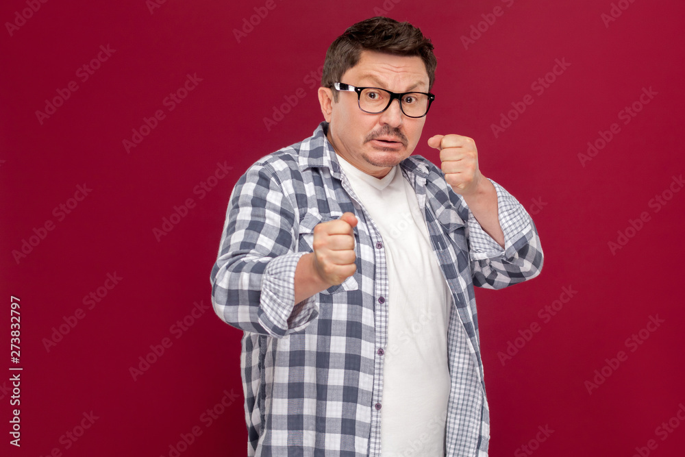 Portrait of angry handsome middle aged business man in casual checkered shirt and eyeglasses standing with boxing fists and ready to attack. indoor studio shot, isolated on dark red background. Portrait of angry handsome middle aged business man in casual checkered shirt and eyeglasses standing with boxing fists and ready to attack. indoor studio shot, isolated on dark red background.