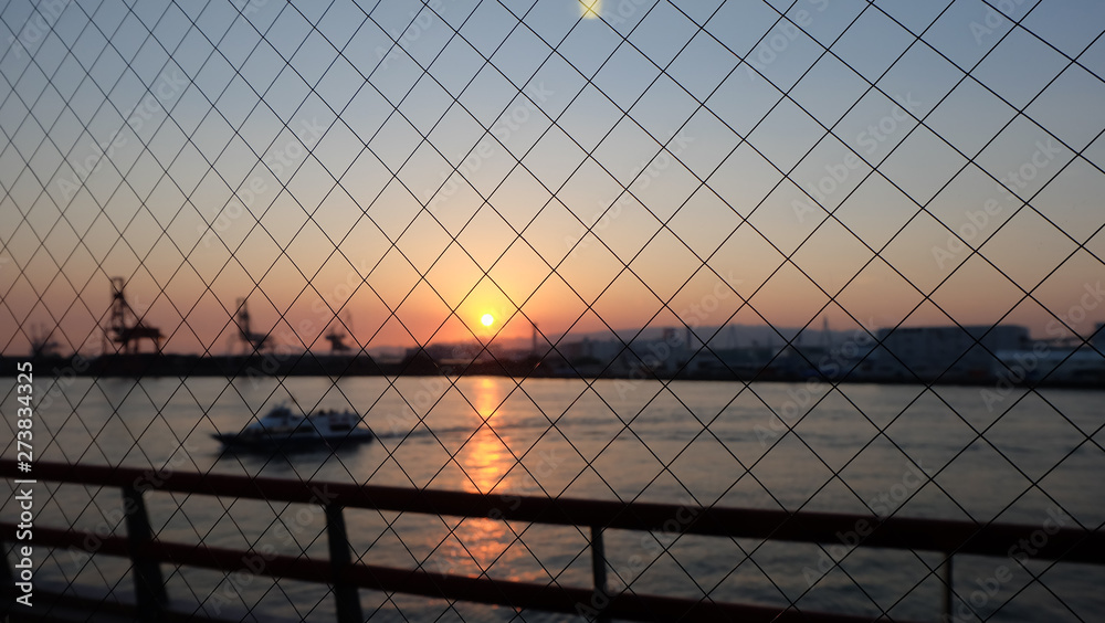 Fototapeta Wire fence, with blurred background of sea with ship, outlines of port cargo cranes and buildings, against a setting sun with twilight in the sky,