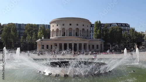 Paris, France - April 5, 2019: Rotonde de La Villette with fountain in the foreground and lots of people walking down the street. Blue sky background. photo